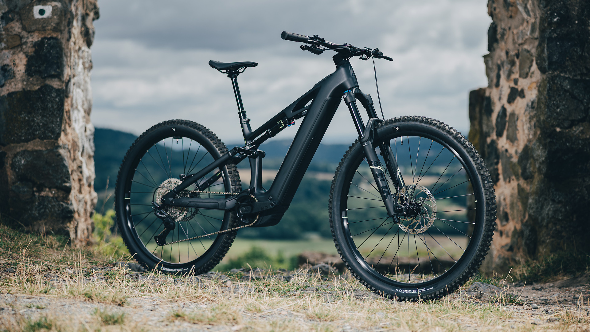 Električno gorsko kolo CUBE Stereo Hybrid ONE44 HPC Race na travi ob kamniti ograji z razgledom na dolino v ozadju